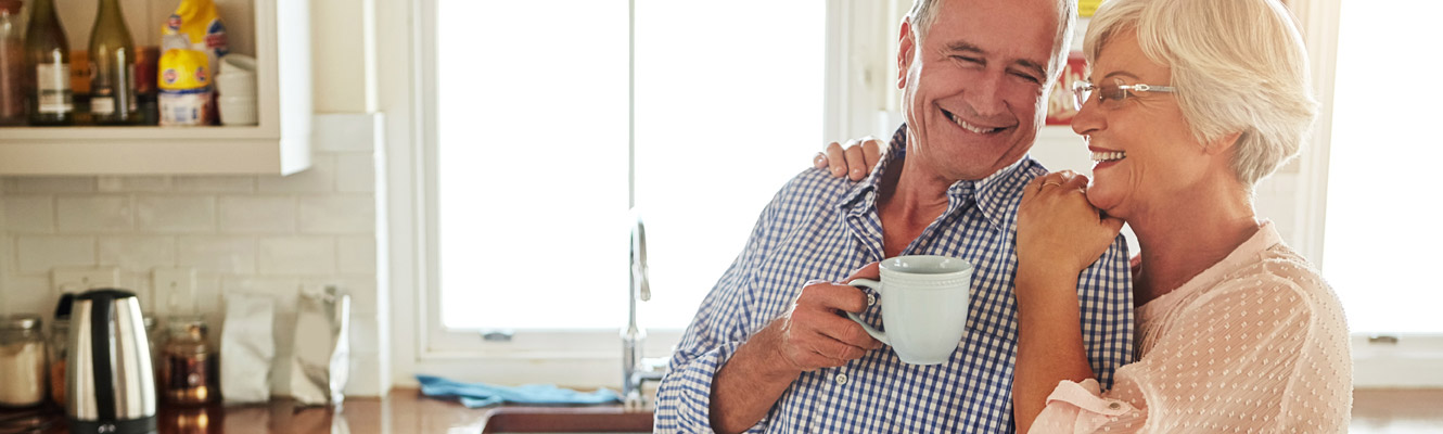 An elderly couple smiles as they hug in their kitchen and enjoy coffee.